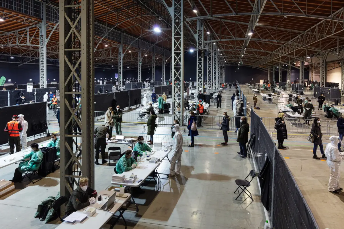Mass vaccination center in a large industrial hall; workers in protective suits sit at long tables with medical supplies amid metal framework.