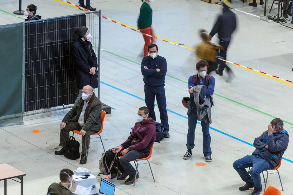 Indoor hall with masked individuals seated on orange chairs; a metal barrier on the left and people walking by.