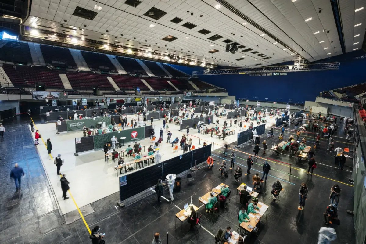 Mass vaccination center in indoor arena; people sit at desks behind partitioned stations.