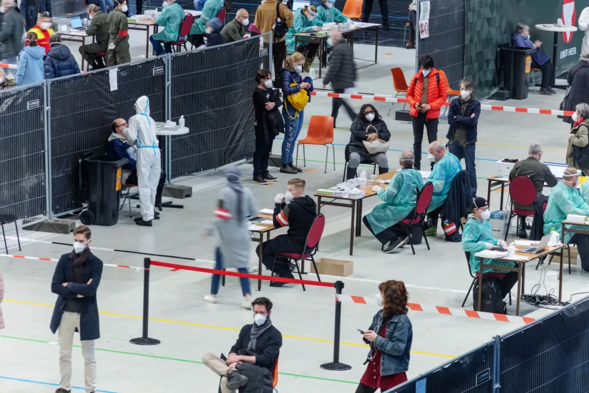 Vaccination clinic set up in a gymnasium; healthcare workers in PPE administer vaccines while people in masks wait behind barriers.