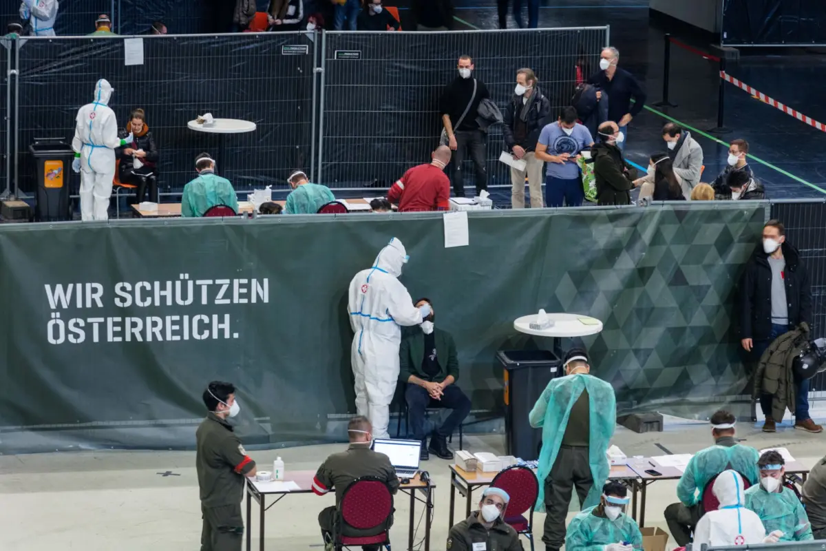 Medical staff in protective suits and masks conduct indoor COVID-19 testing; people queue behind metal fencing.
