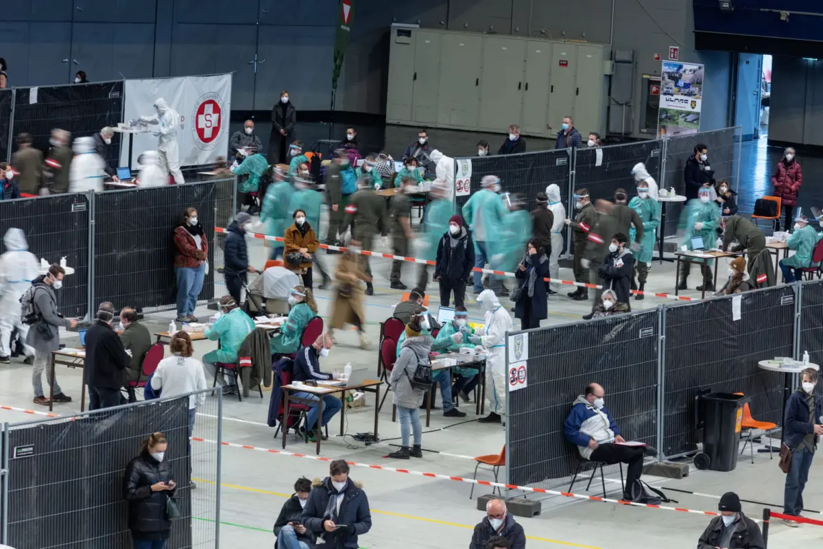 Healthcare workers in full PPE administer vaccines to seated people at a large indoor vaccination center.
