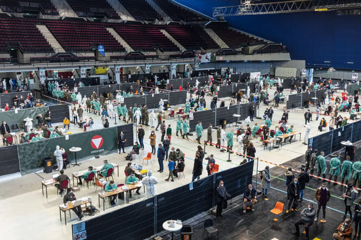 Mass vaccination site inside arena; healthcare workers in PPE vaccinating and guiding seated and standing attendees.