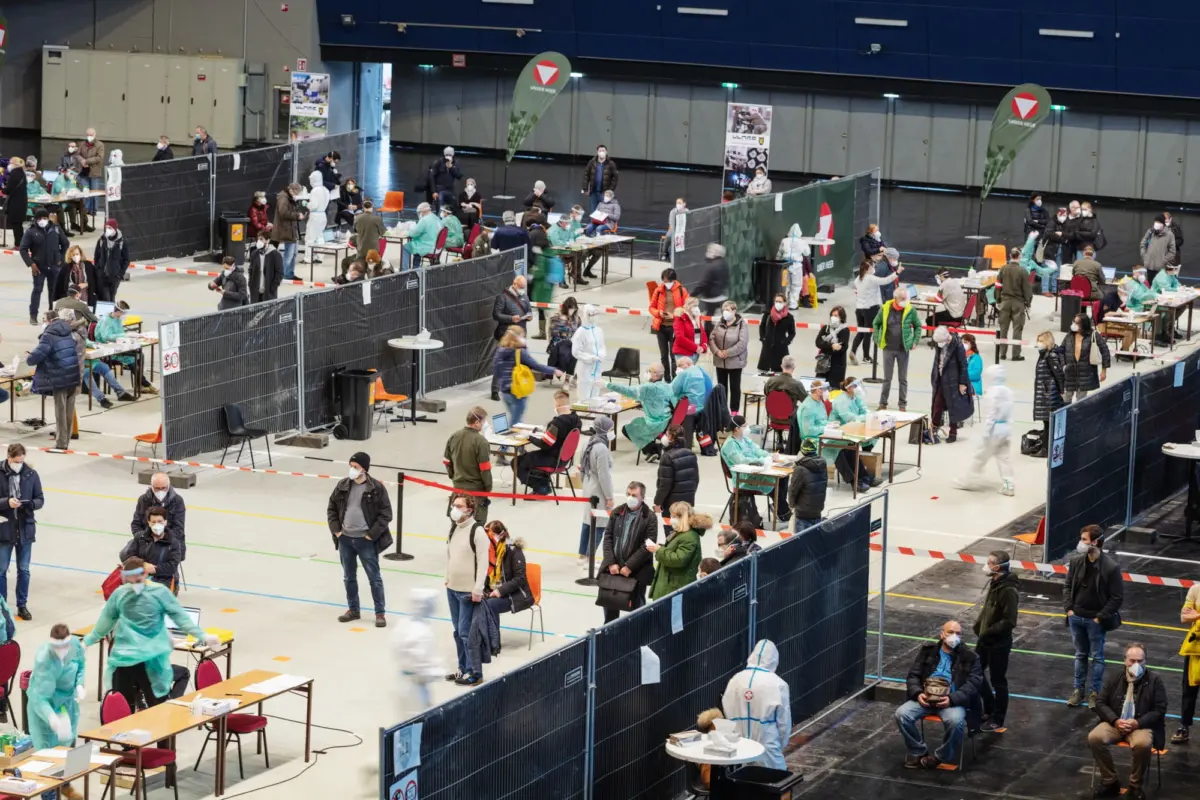 Mass vaccination clinic in a gymnasium; healthcare workers in protective suits administer vaccines to seated and standing attendees.