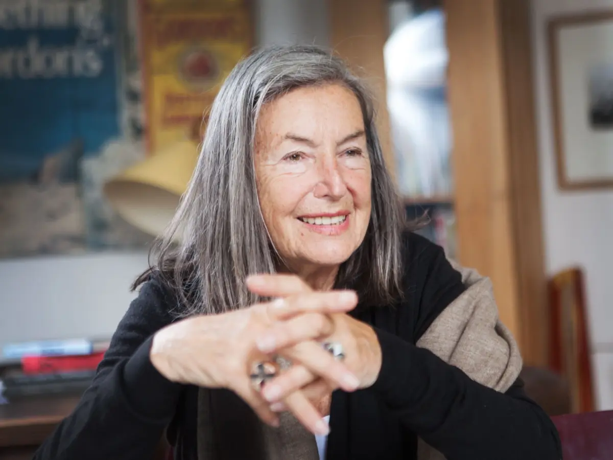Older person with long gray hair smiles while gesturing with hands indoors, in a room with shelves and posters in the background.