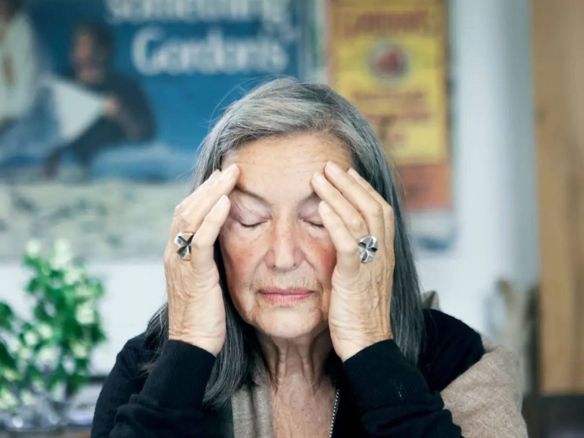 Elderly woman with gray hair presses her temples with fingertips, eyes closed, indoors.