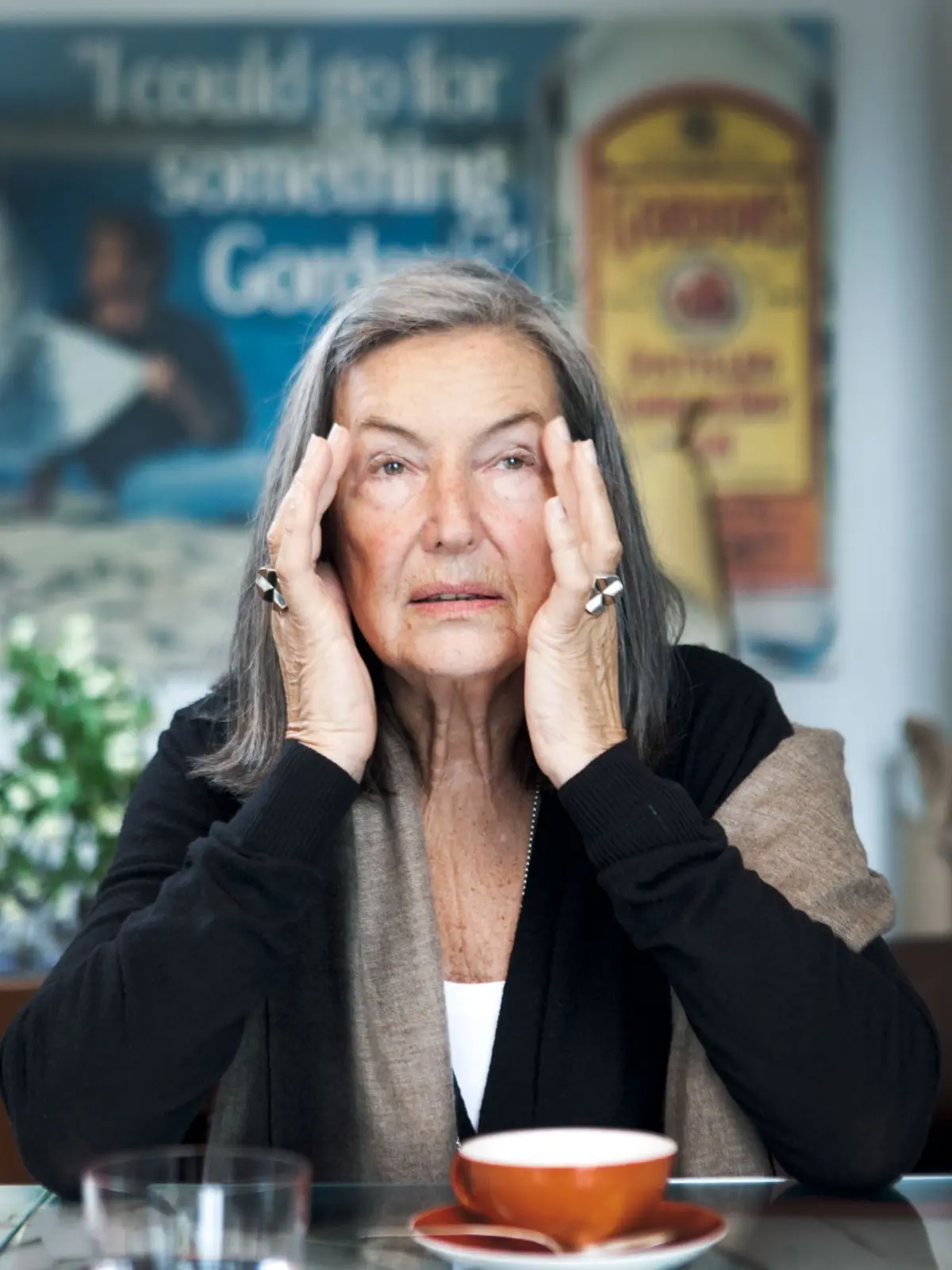 Senior woman with gray hair presses temples while seated at a cafe table with an orange cup.