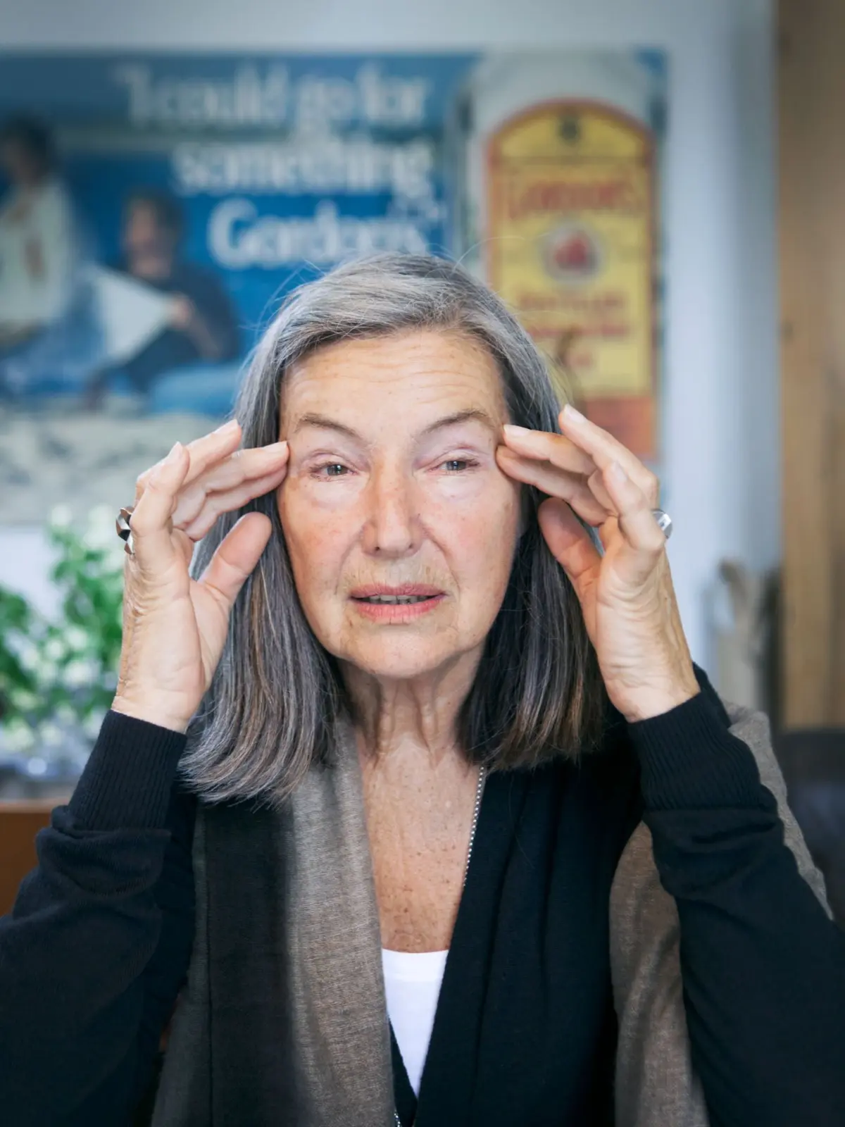 Older adult with gray hair rubs temples with fingertips indoors; blurred posters in the background.