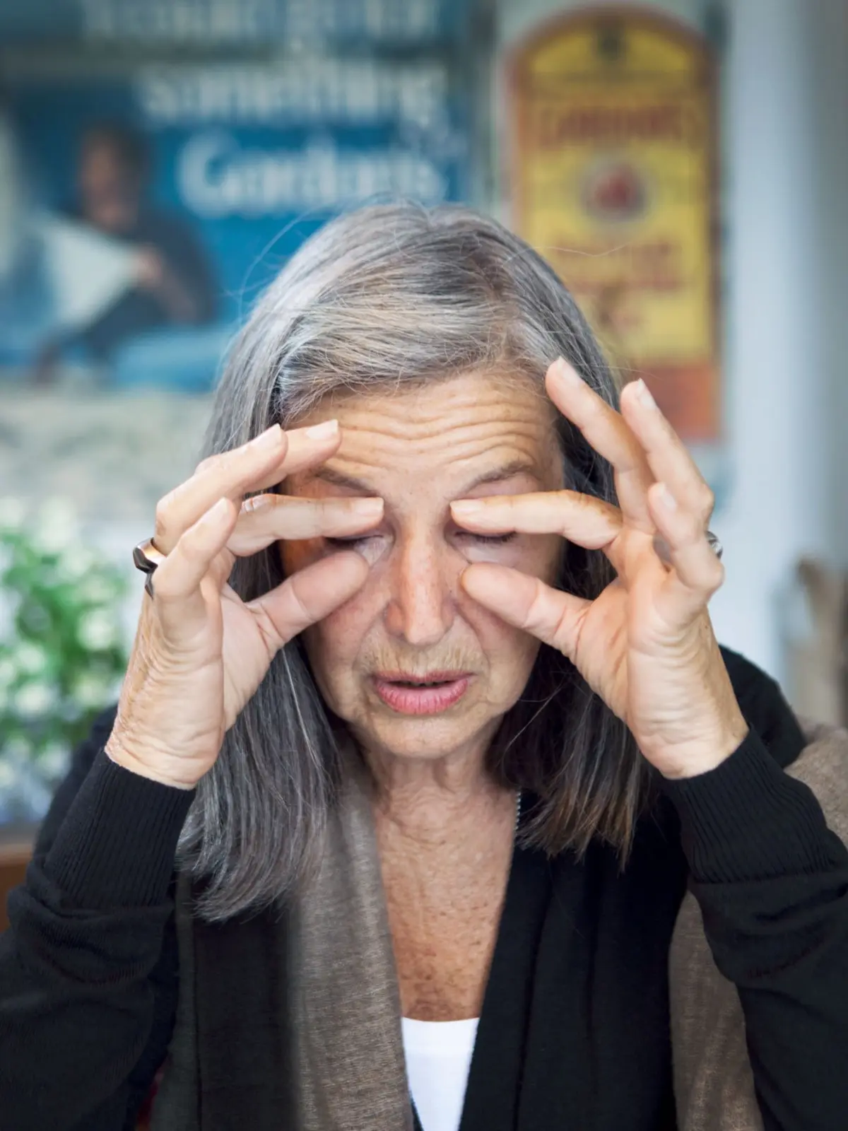 Elderly woman with gray hair presses temples with her fingers, eyes closed, in front of blurred posters.