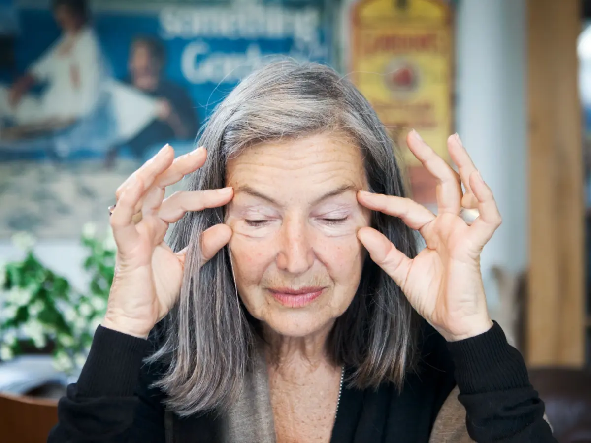 Older person with gray hair closes eyes, pressing fingertips to temples in an indoor setting.