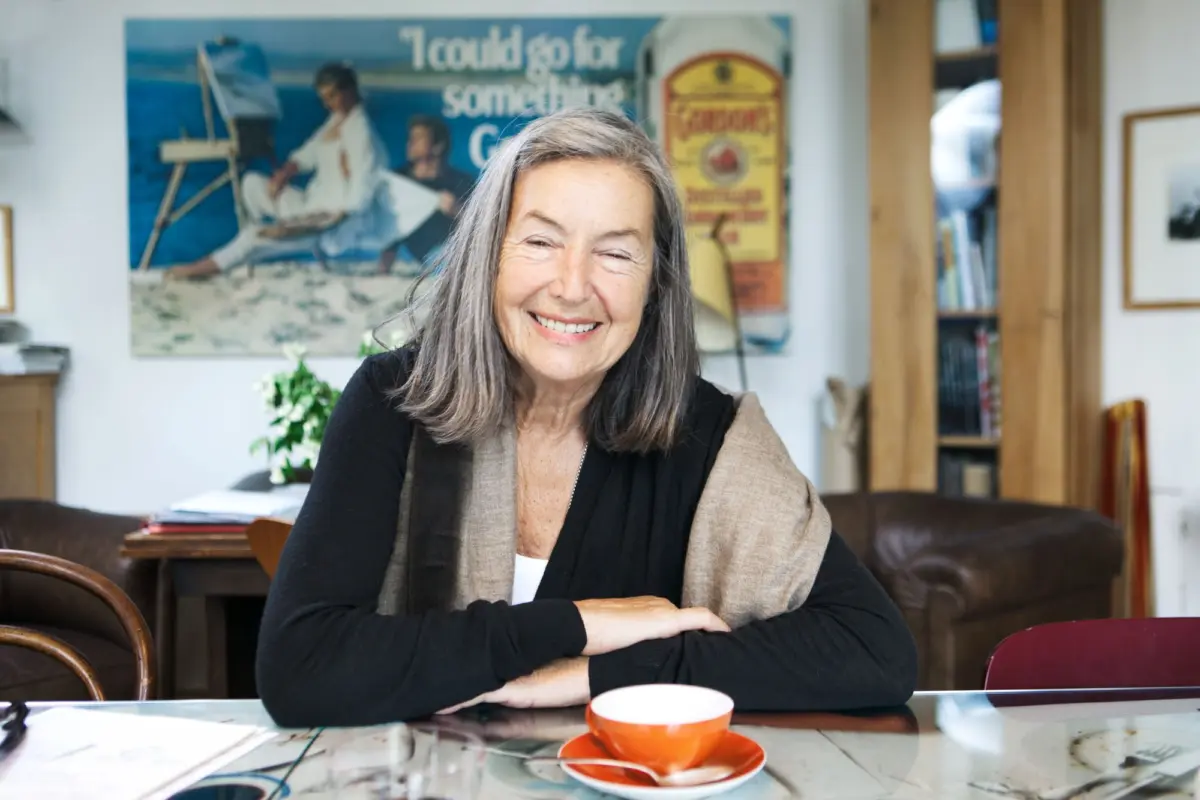 Older adult with gray hair smiles, seated at a glass table with arms folded in a living room with a bookshelf and artwork in the background.