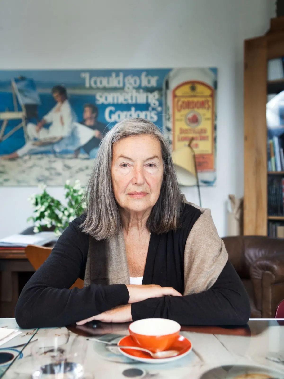Older woman with gray hair sits at a glass table, arms folded, in a room with posters and a bookshelf.