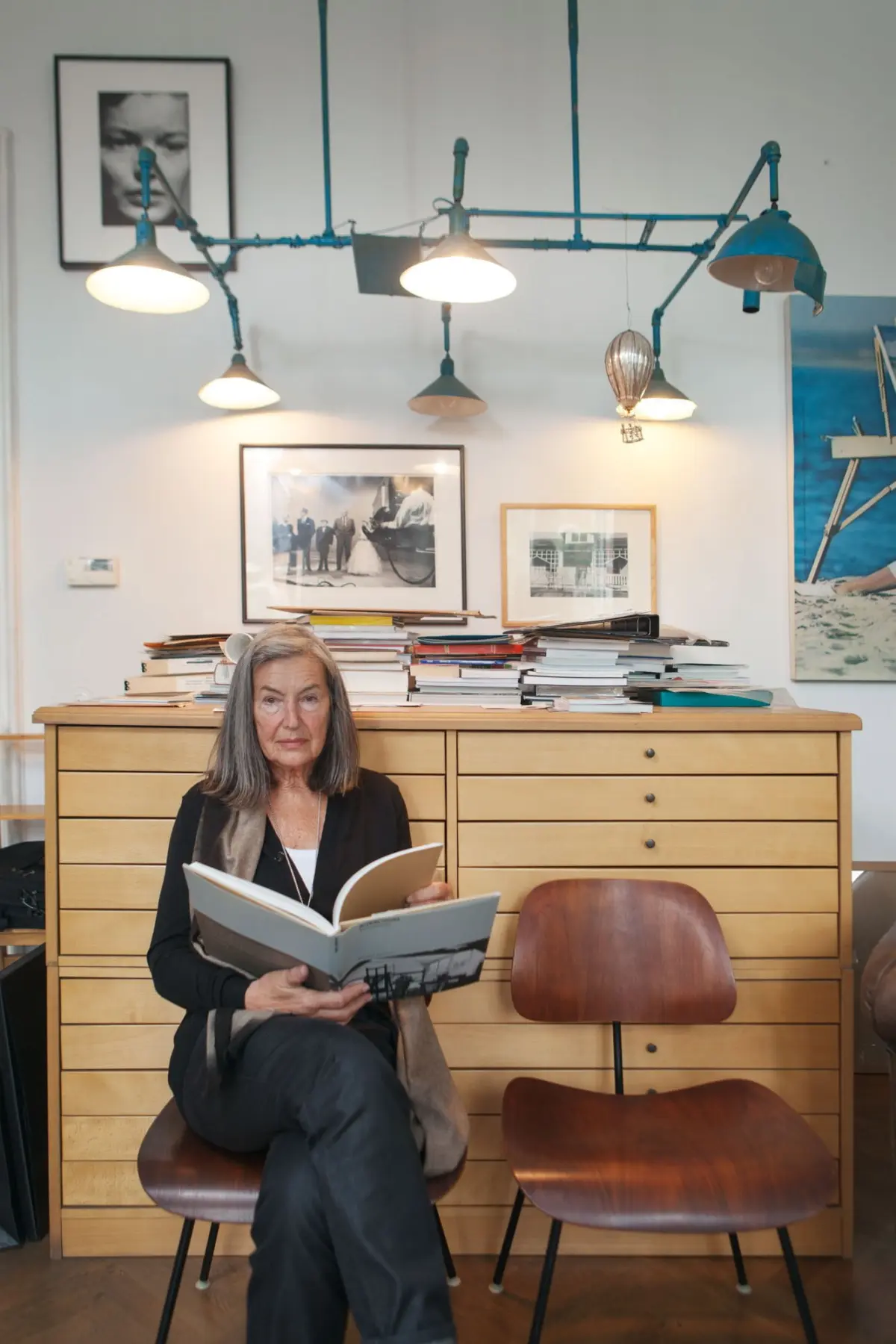 Older person seated on a wooden chair, reading an open magazine amid a dresser stacked with books and framed photographs in a cluttered room.