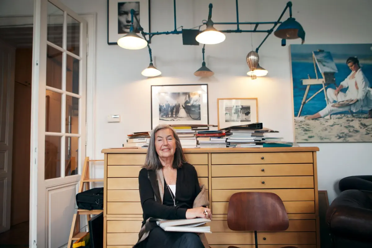 Older woman sitting at a wooden dresser stacked with papers, smiling, in a home office with framed photos and a beach poster on the wall.