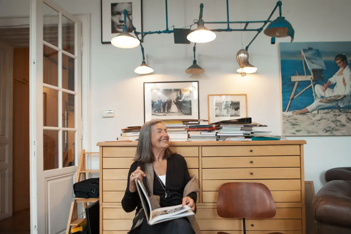 Older woman with gray hair, smiling while flipping through a magazine, seated in a cluttered home office with a wooden dresser.
