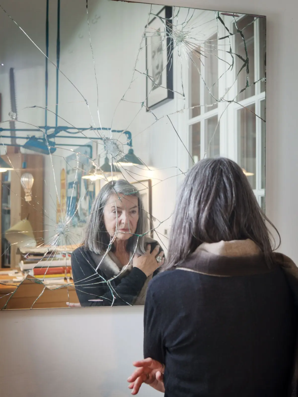 Shattered mirror shows two people in an office; gray-haired person faces a dark-haired person, with a desk and lamp visible.