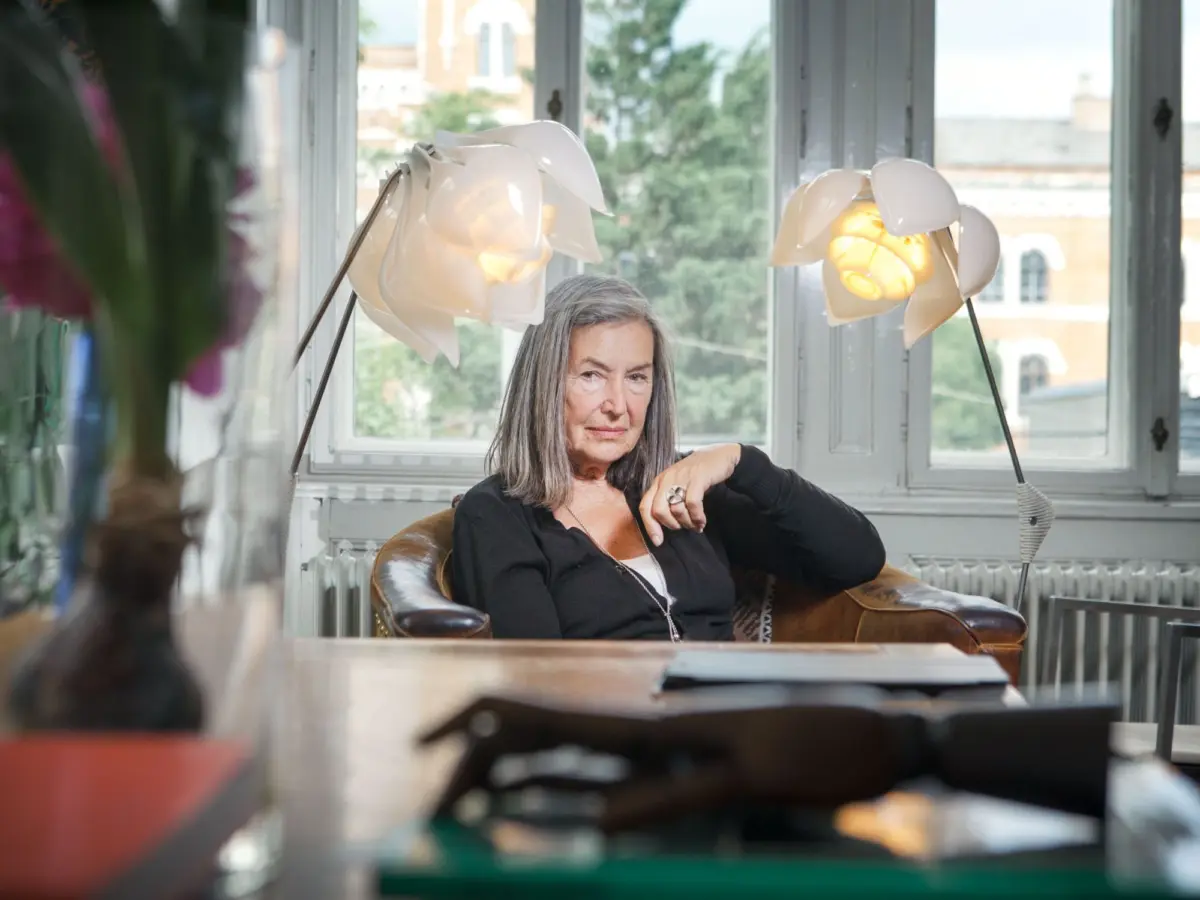 Older woman with gray hair sits in a brown leather chair by a window, near white flower-shaped lamps.