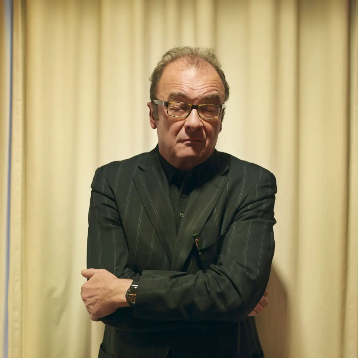 Middle-aged man in a pinstripe suit with glasses, arms crossed, standing indoors in front of beige curtains.