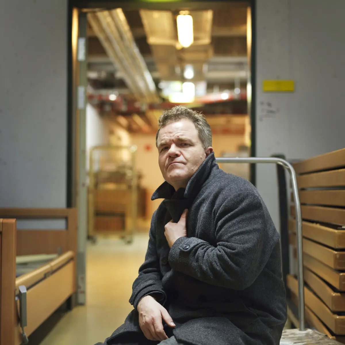 Adult in dark wool overcoat sits on a bench in a warehouse corridor, adjusting coat collar.