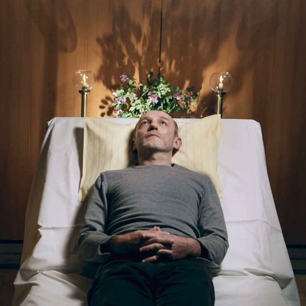 Adult person lies supine in bed, hands clasped over abdomen, head on pillow; floral arrangement and lit lamps against wood-paneled wall.