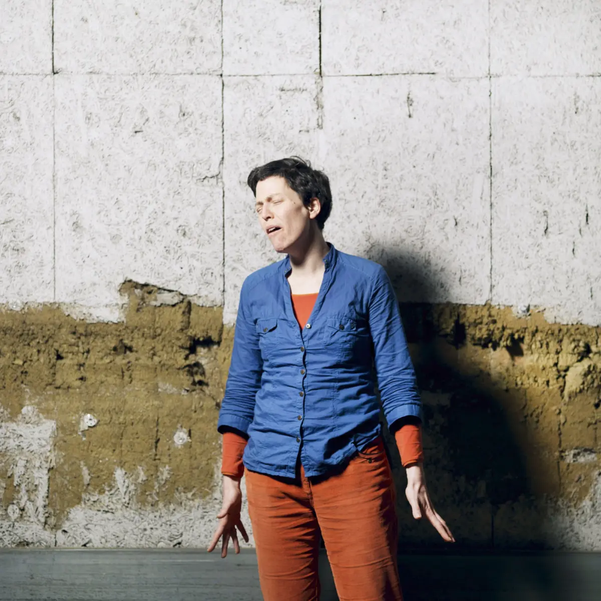 Woman in a blue button-up shirt and orange pants sings with eyes closed beside a weathered white wall.