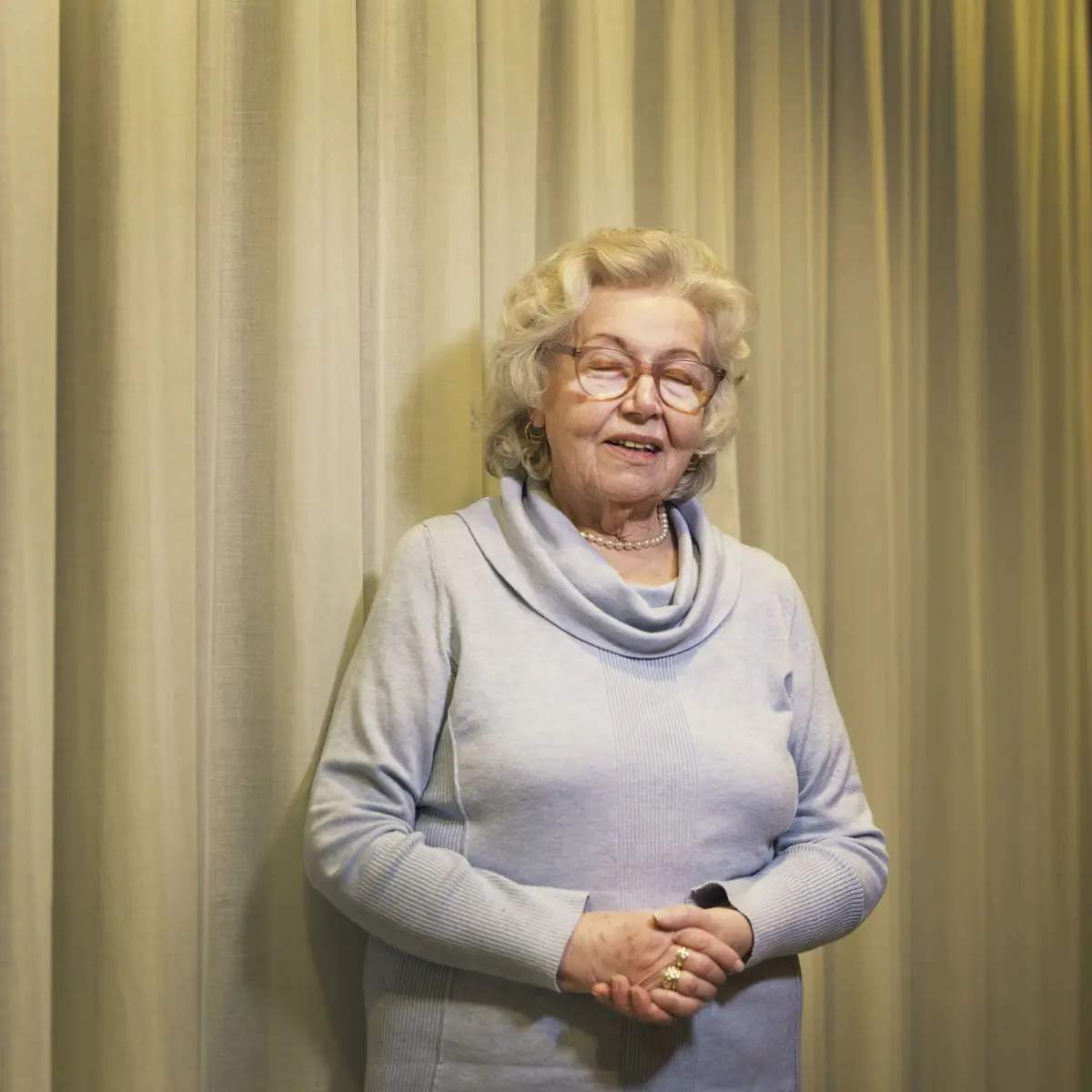 Elderly woman with glasses, smiling, wearing a light blue sweater and scarf; hands clasped in front, standing before beige curtains.