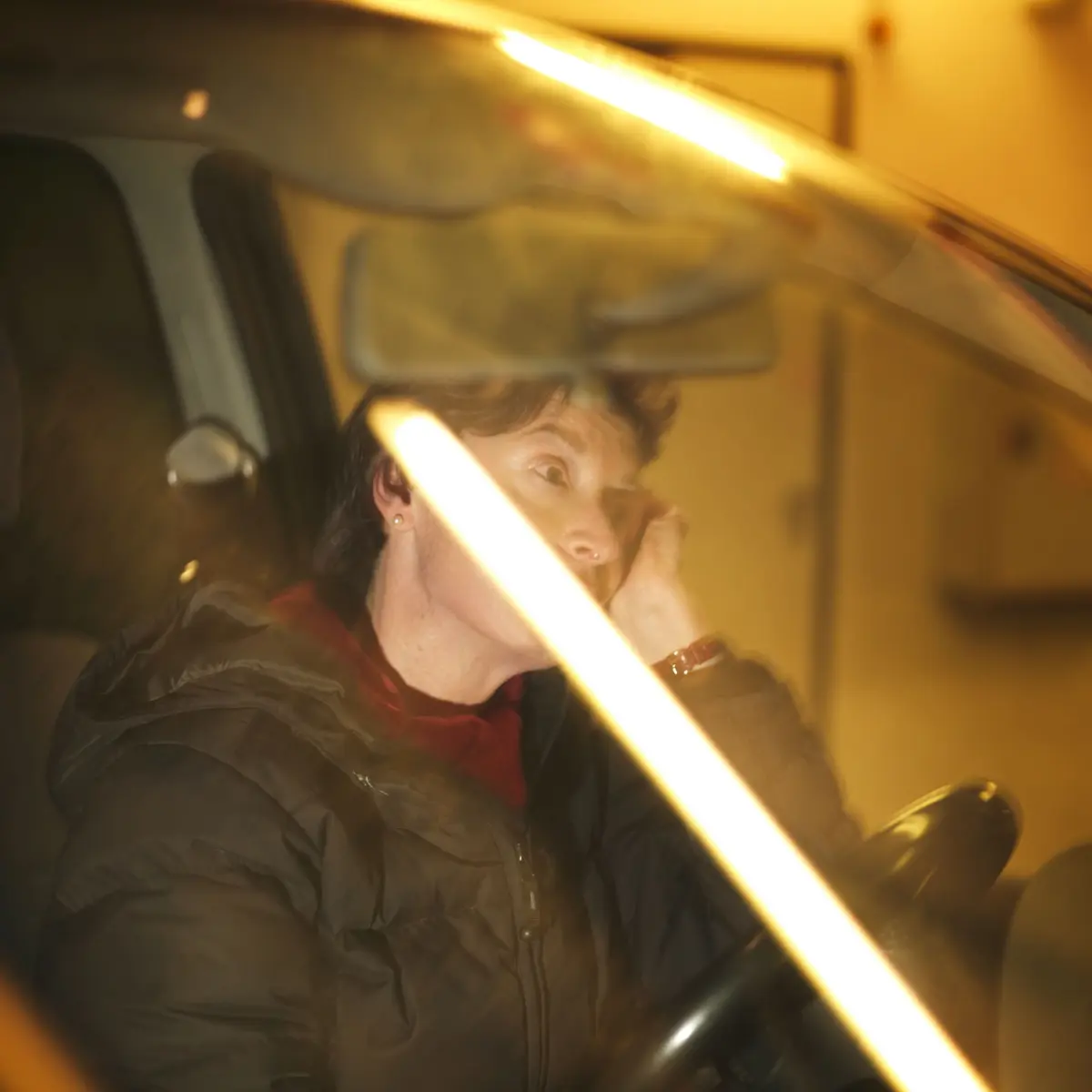 Person in a dark puffer jacket sits in the driver's seat of a car, resting their head on their hand in a dim garage.