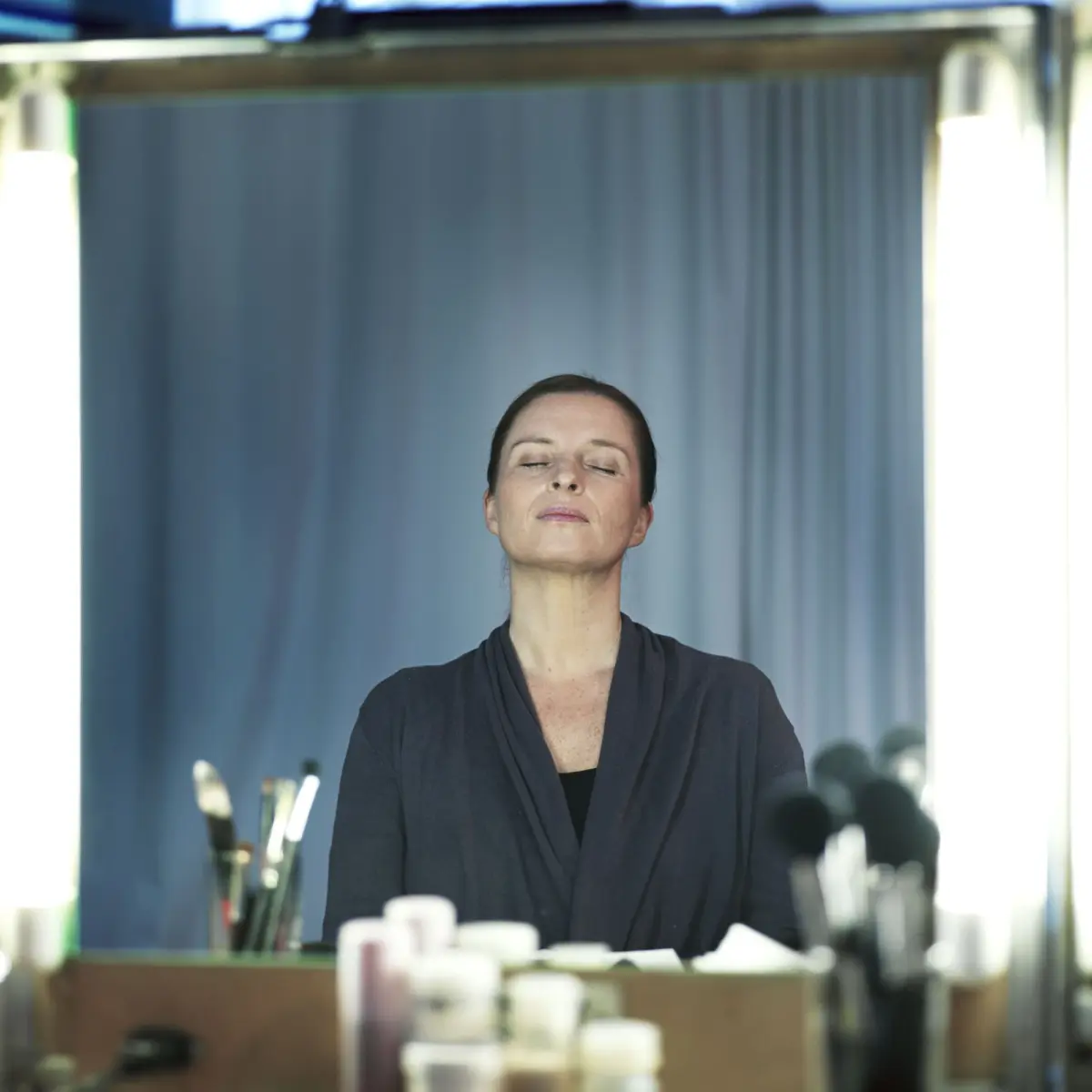 Person with eyes closed wearing dark robe, seated at makeup vanity; cosmetics and brushes on the table, blue curtain backdrop.