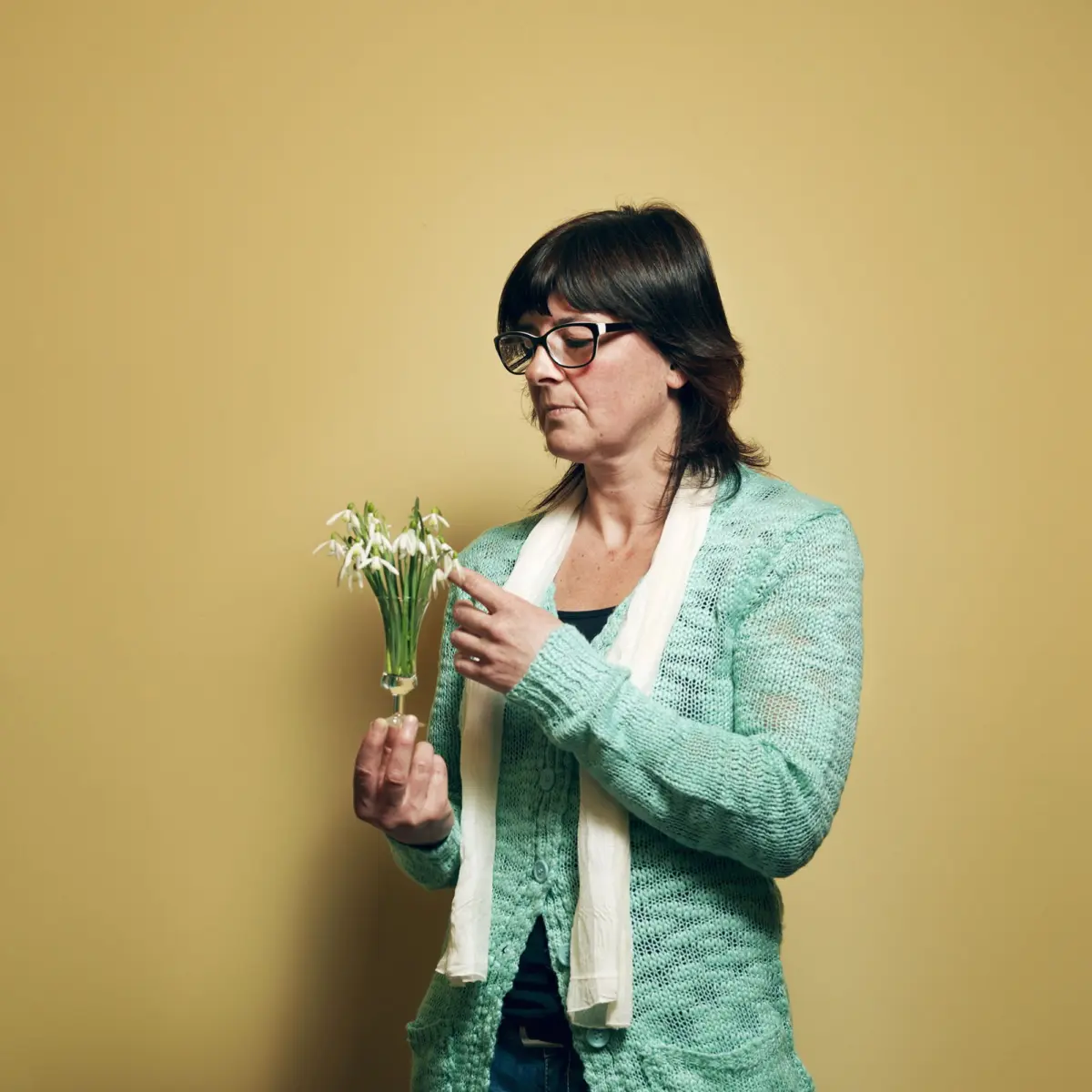 Adult person with glasses in a mint cardigan and white scarf holds a bouquet of snowdrops and points at it against a yellow studio backdrop.
