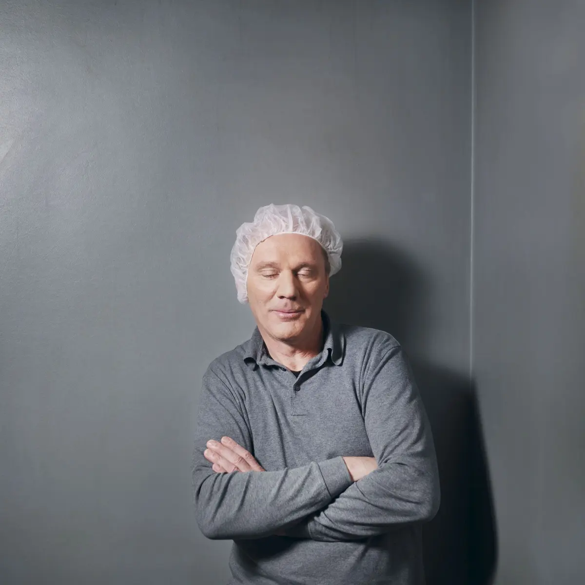 Man wearing a disposable hairnet and gray polo, standing with arms crossed and eyes closed against a gray wall.