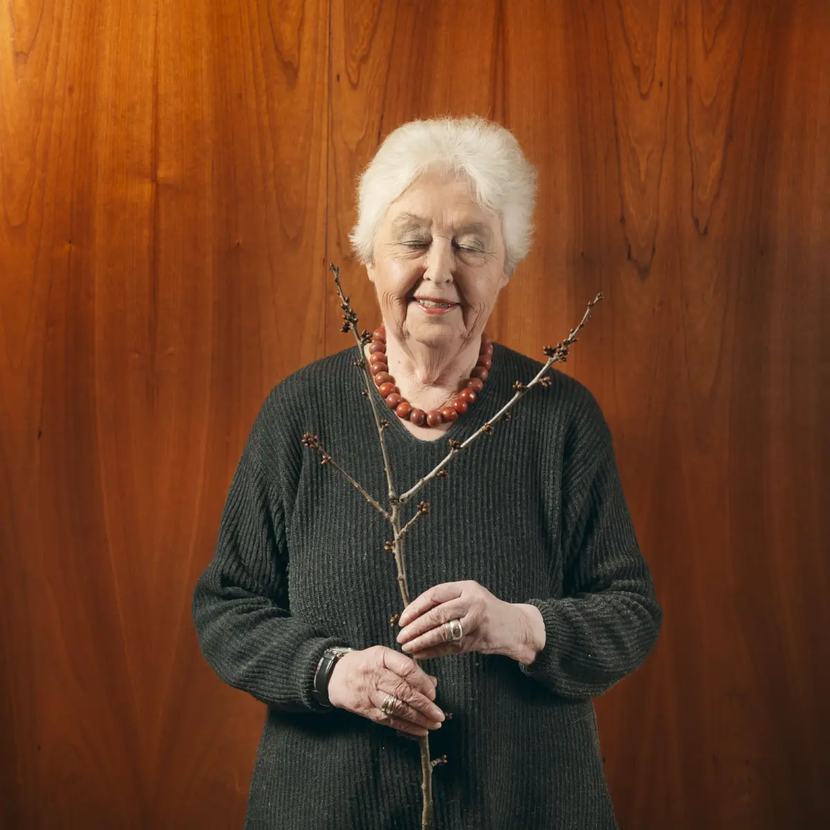 Older woman with white hair and a beaded necklace smiles with eyes closed, holding bare branches against a wood-paneled wall.