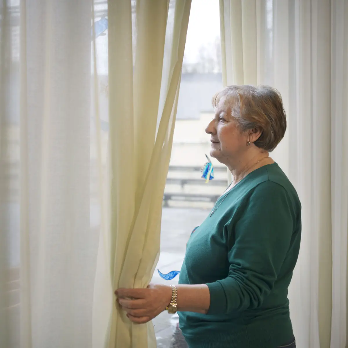 Elderly woman in a green sweater stands indoors, holding a cream curtain and looking out the window.