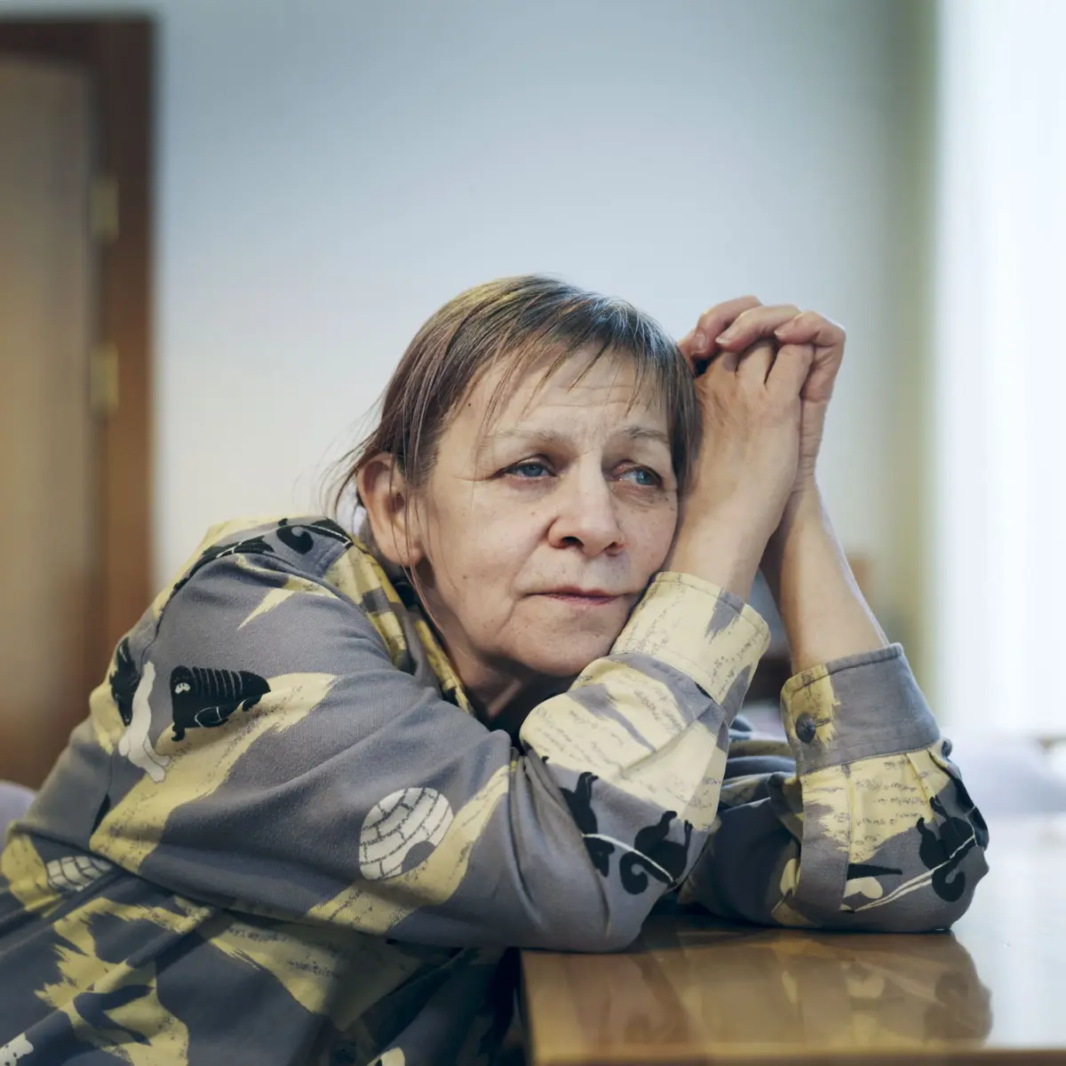 Older woman indoors, leaning on a wooden table with clasped hands, wearing a camouflage-patterned shirt, appearing pensive.