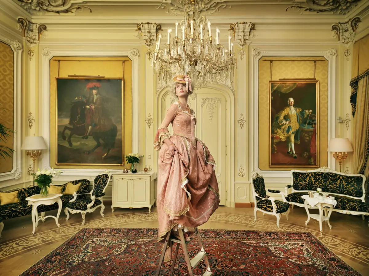 Person in pink rococo gown stands on a stepladder beneath a crystal chandelier in an opulent room with gilded walls and grand portraits.