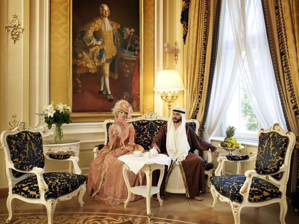 Two adults in 18th-century dress at a tea table in a gilded parlor; peach gown with fan beside robe-clad companion, large portrait behind.