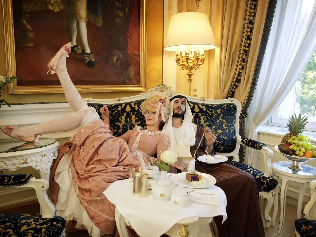Two people in 19th-century attire lounge on a gilded blue-and-gold sofa in an ornate parlor; one person's leg rests on an ottoman.