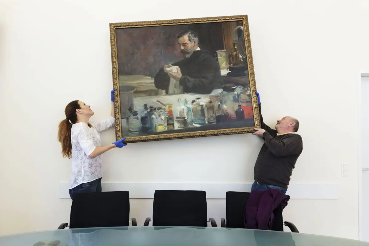 Two people lift a large framed painting depicting a bearded man in a studio with bottles on a table.
