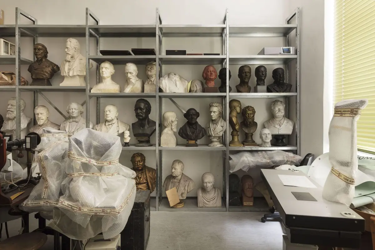 Art studio stocked with plaster busts on metal shelves; wrapped sculpture on a chair in foreground and a workstation to the right.