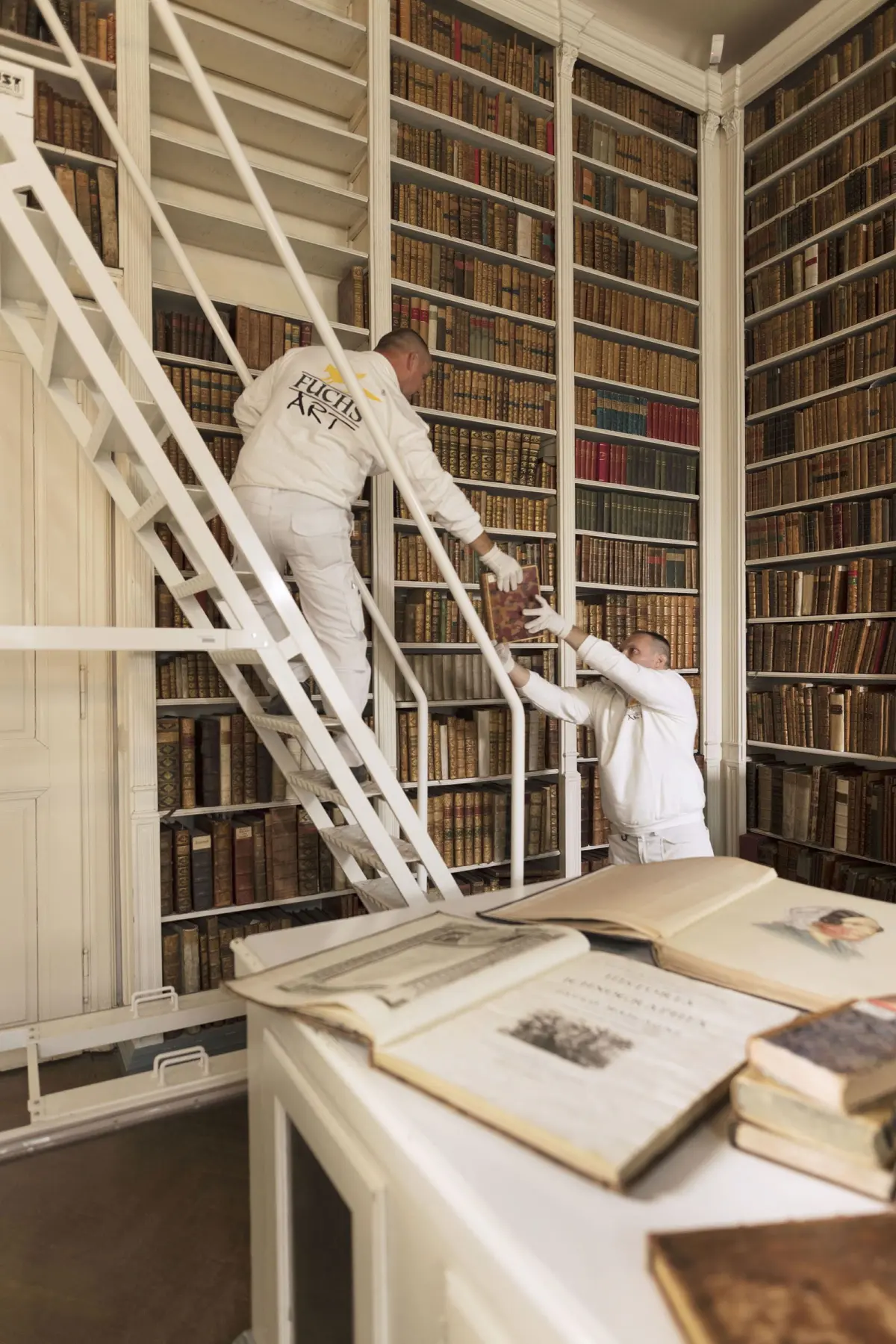 Two workers in white uniforms pass a book up a white metal staircase in a tall, antique library filled with leather-bound volumes.