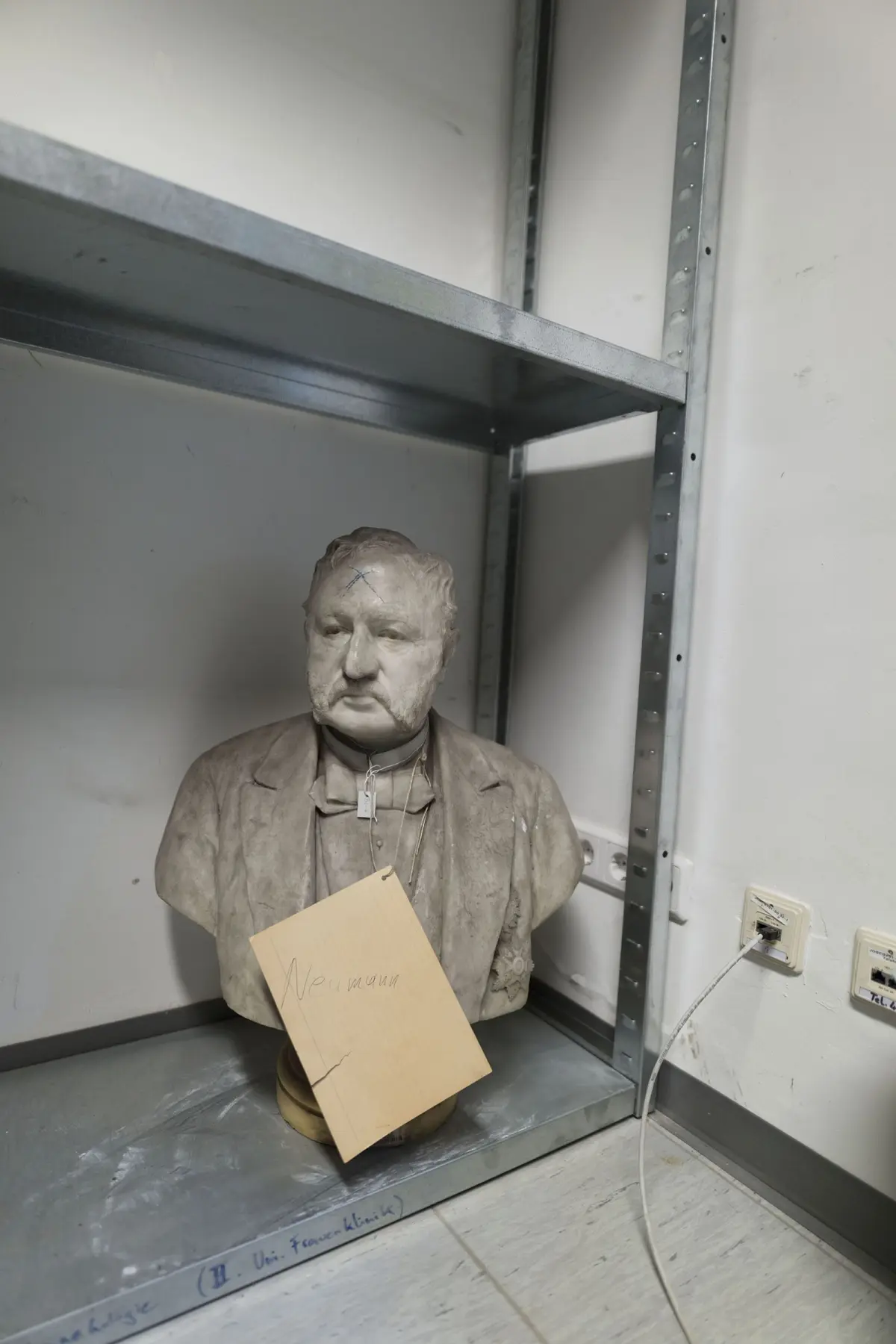 Stone bust of a person in a suit on a metal shelf in a storage room, with a hanging tag.