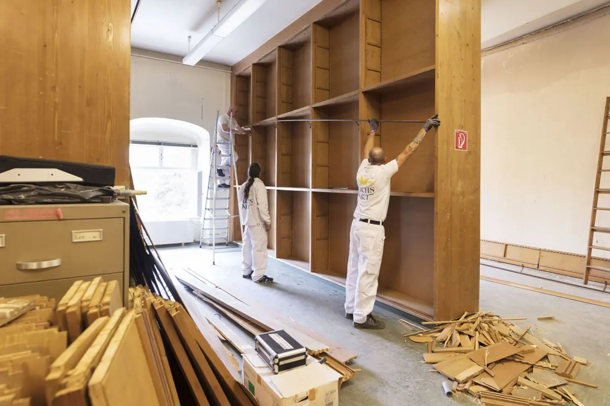 Three carpenters in white coveralls install a large wooden shelving unit in a workshop.