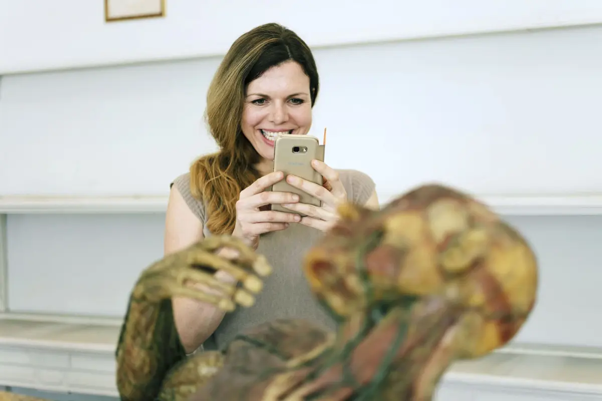 Smiling woman uses smartphone to snap a photo of a zombie prop in a bright classroom.