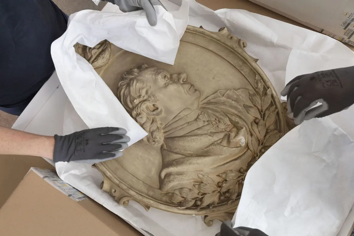 Circular plaster bas-relief depicting a male profile with laurel and drapery, being unwrapped by gloved hands.