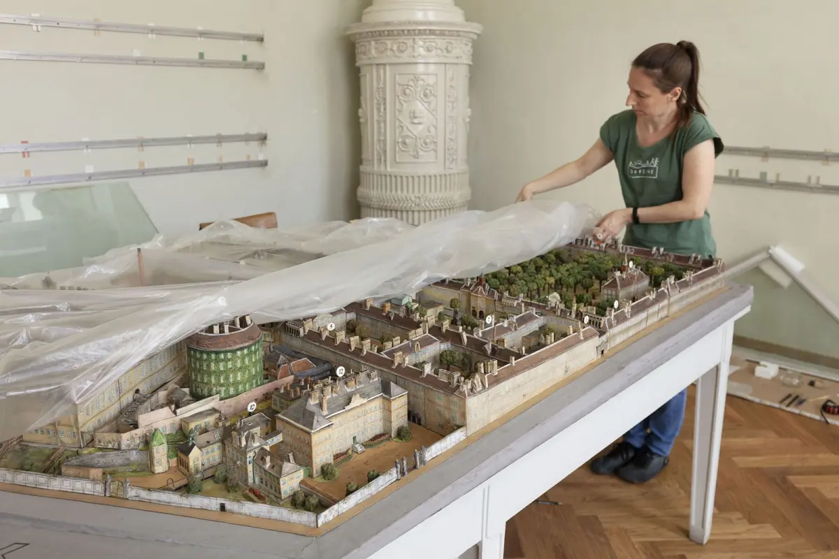 Woman in a green shirt adjusts a large-scale miniature town on a table covered with plastic indoors, beside an ornate white column.