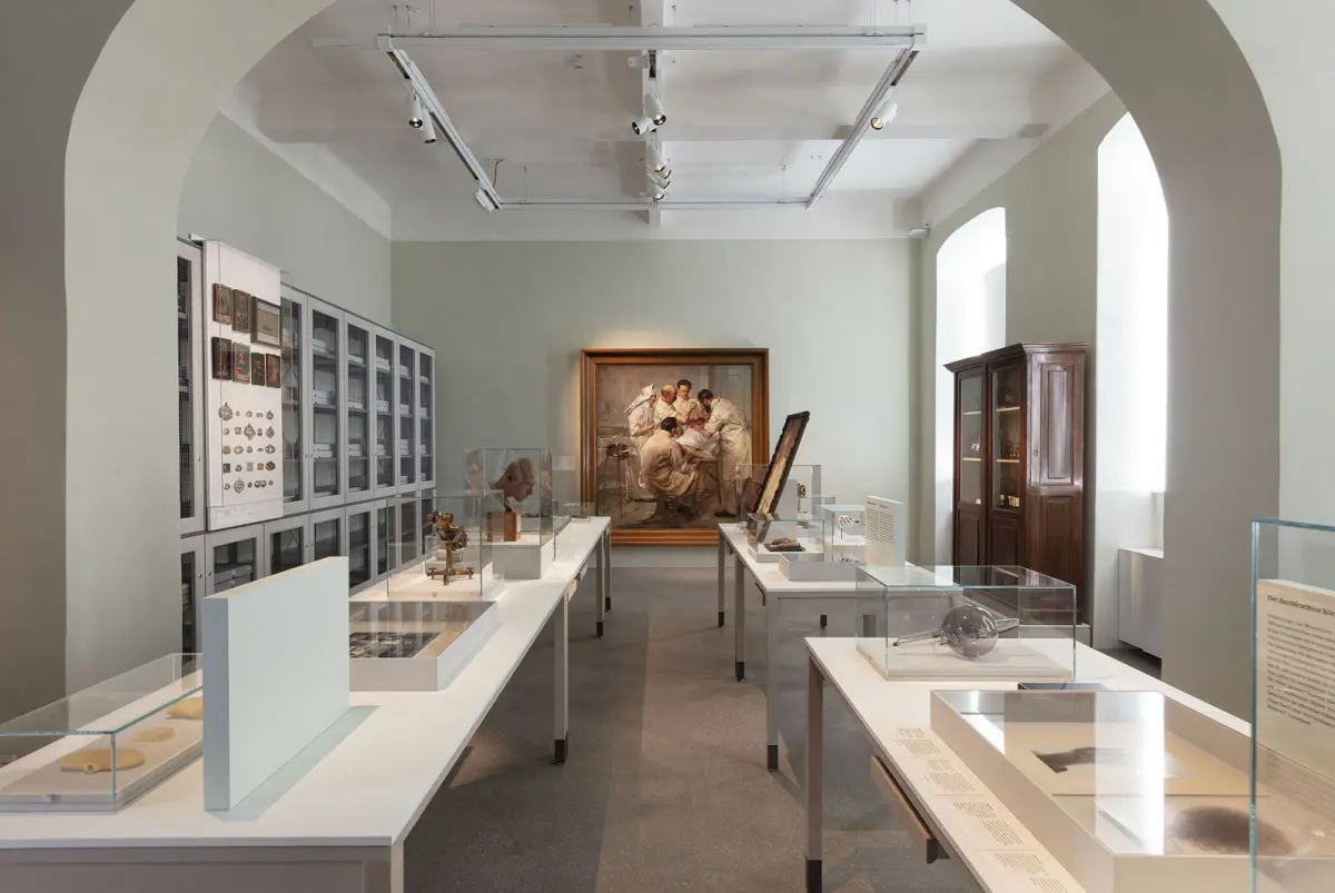 Museum gallery with glass display cases, artifacts on tables, and a large framed painting at the rear; a wooden cabinet by the window.