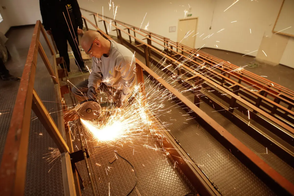 Bald worker wearing safety glasses grinds metal; sparks erupt in a workshop with orange rails.