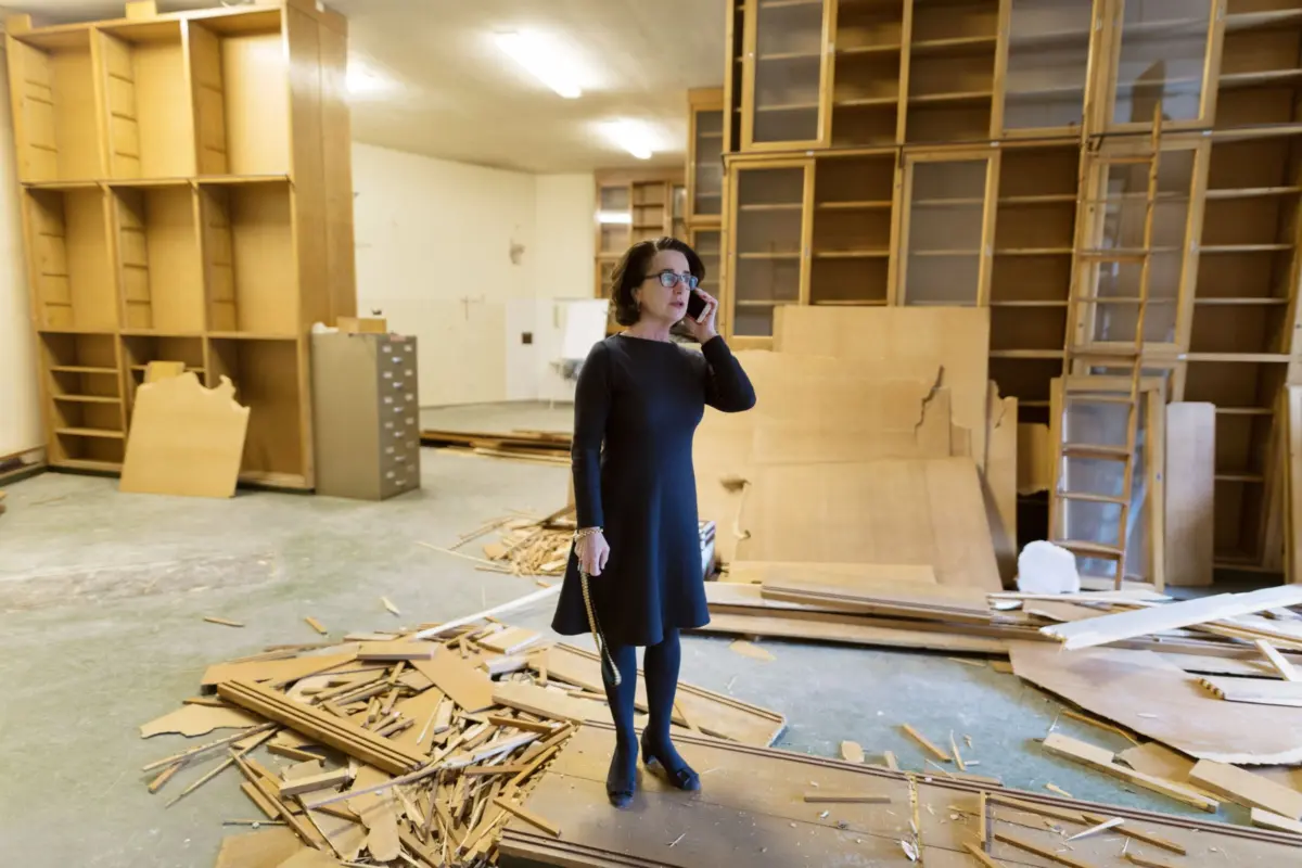 Woman in a black dress talking on a cellphone in a cluttered woodshop with tall wooden shelves and scattered plywood pieces.