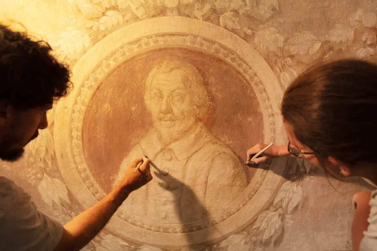 Two artists sketch a circular, weathered mural depicting a human face on a stone wall, using pencils in warm ambient light.