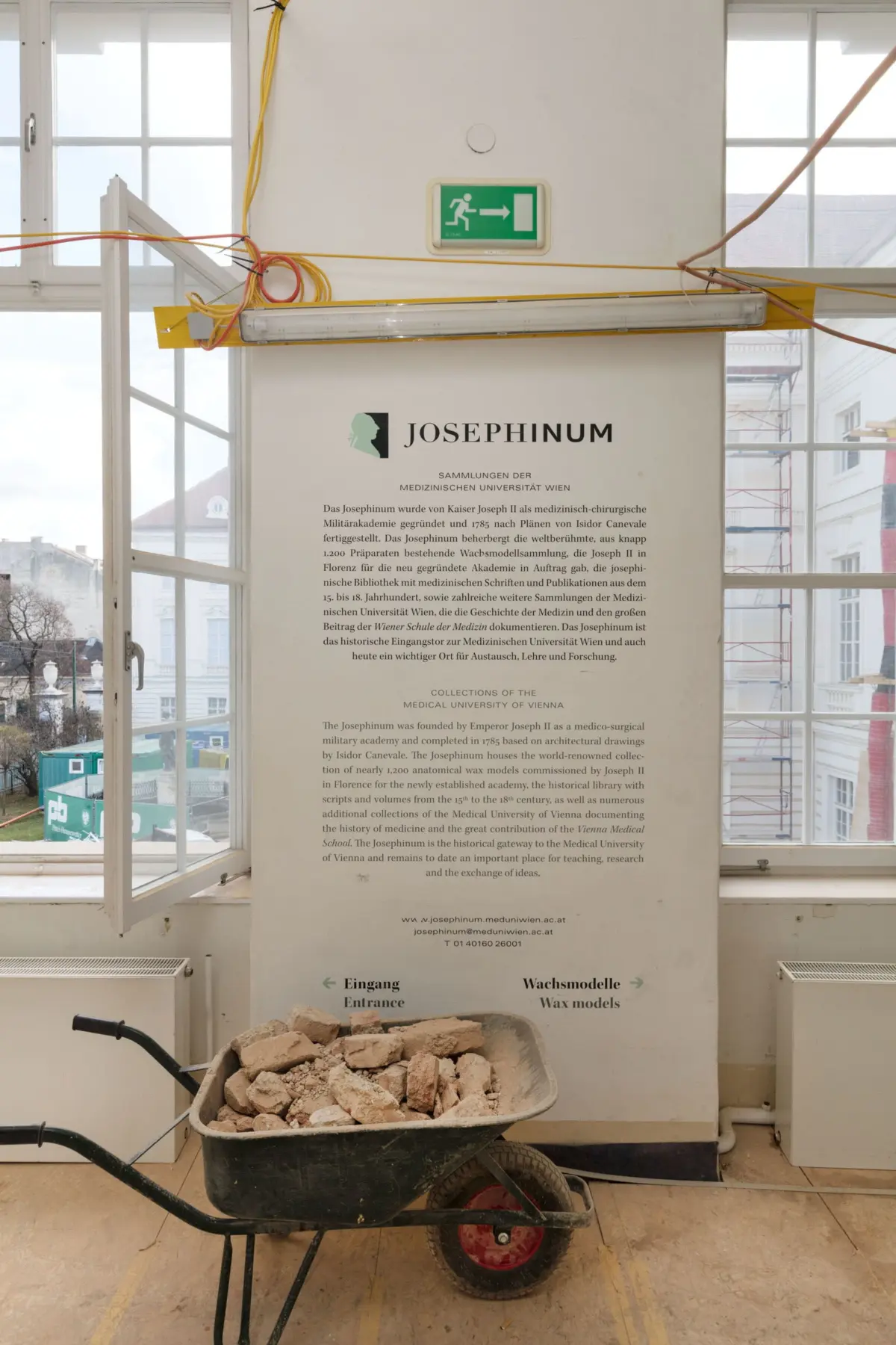 Wheelbarrow full of bricks in a construction area indoors, with large windows and a Josephinum information panel on the wall.