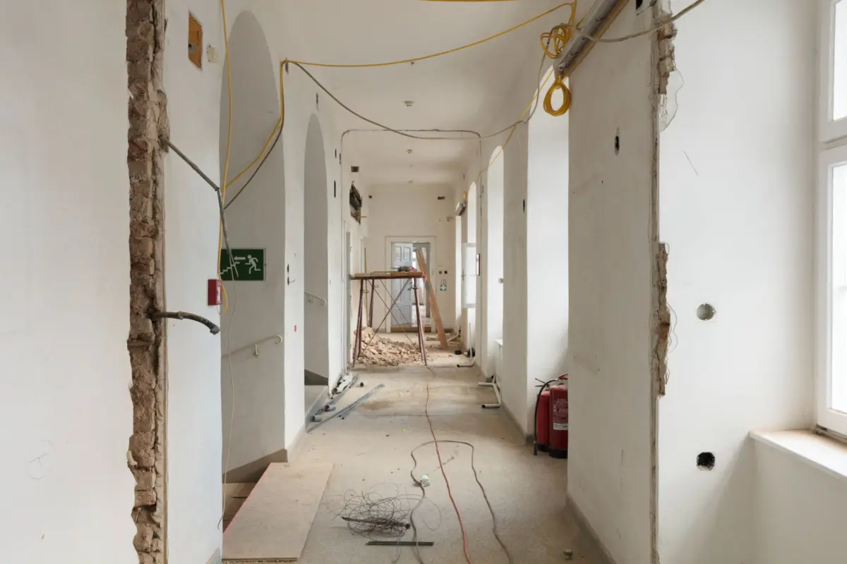 Long hallway under renovation with white walls, exposed brick edges, hanging yellow cables, and scattered debris; scaffold in distance.